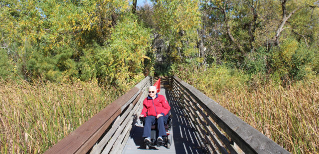 A resident of Regent at Burnsville enjoys a sunny day navigating the scenic Earley Lake Trail in a motorized chair, surrounded by vibrant greenery and peaceful nature.